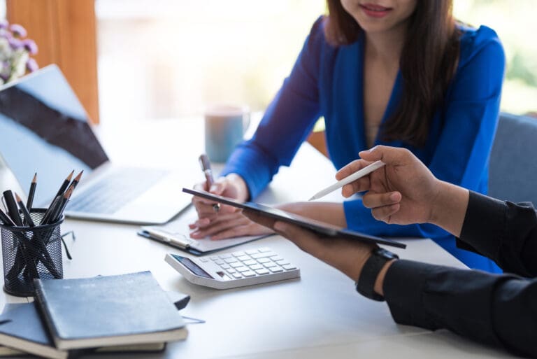 Two business professionals reviewing financial documents with a tablet, calculator, and laptop on a desk.