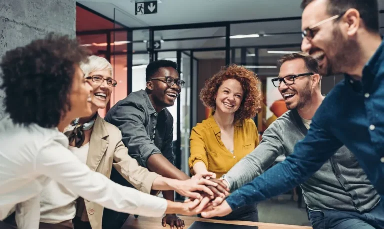 Diverse group of coworkers smiling and stacking hands in a show of teamwork and unity in a modern office setting.