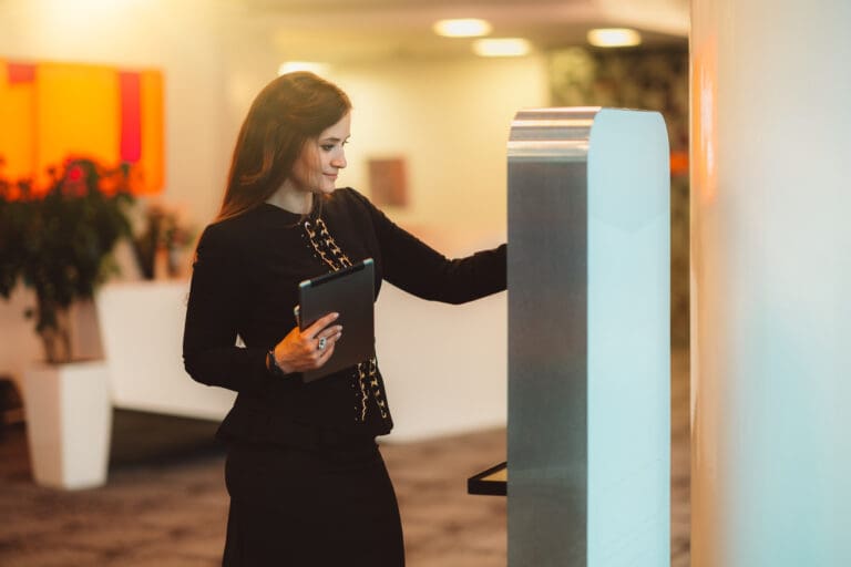 Woman in hotel lobby interacting with a digital kiosk while holding a tablet