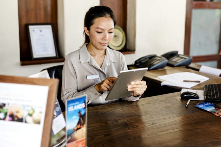 A hospitality professional in uniform using a tablet at a hotel front desk.