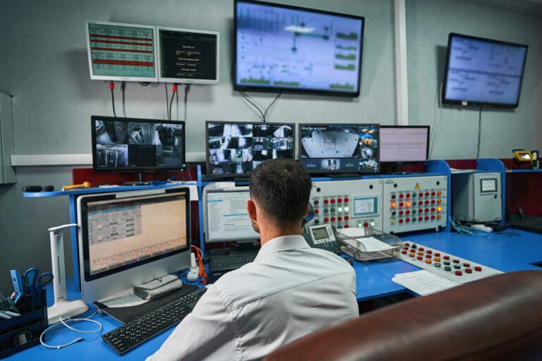 A technology administrator managing multiple monitors and control systems, illustrating the complexity of overseeing interconnected enterprise technology infrastructure.